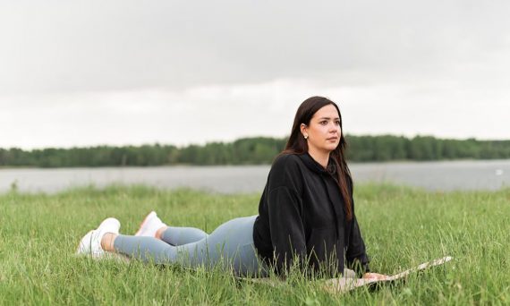 Woman lying on a yoga mat in a grassy field by a lake, promoting relaxation and wellness.